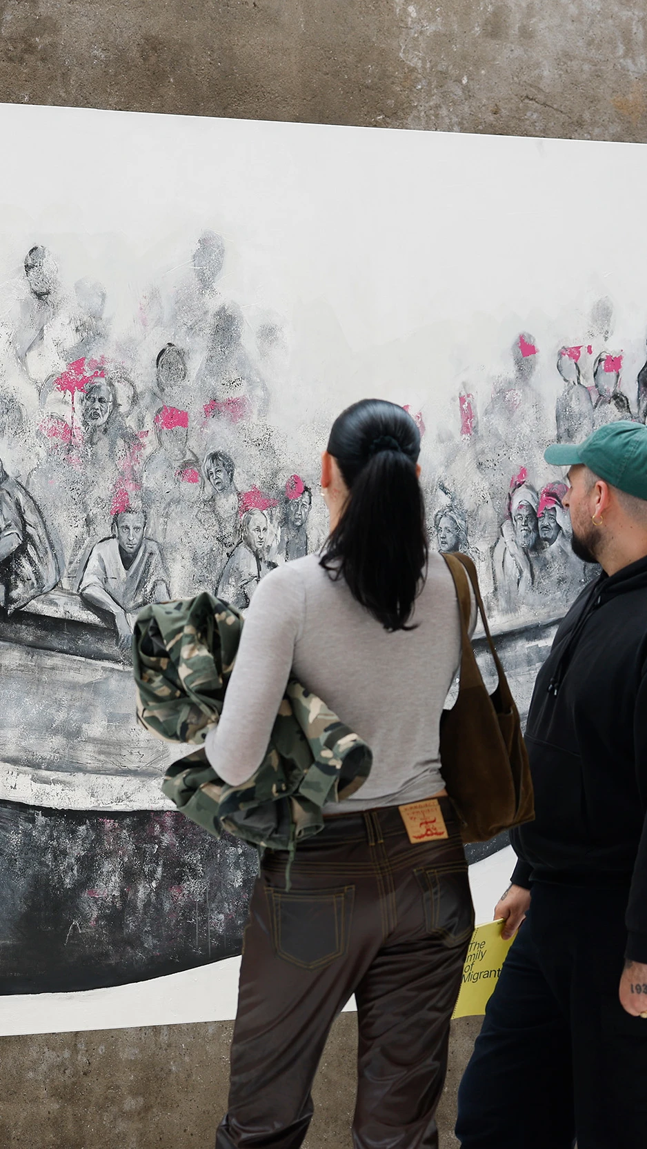 A man and woman observe a large mural of people crowded in a boat, wearing pink life vests. The scen