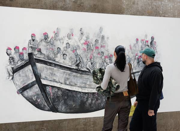 A man and woman observe a large mural of people crowded in a boat, wearing pink life vests. The scen