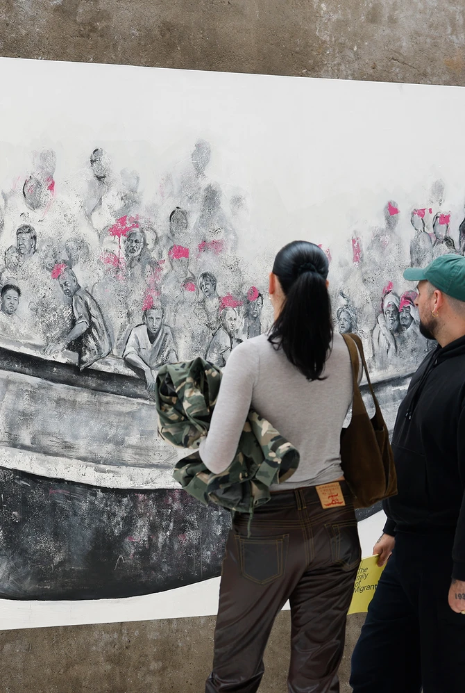 A man and woman observe a large mural of people crowded in a boat, wearing pink life vests. The scen