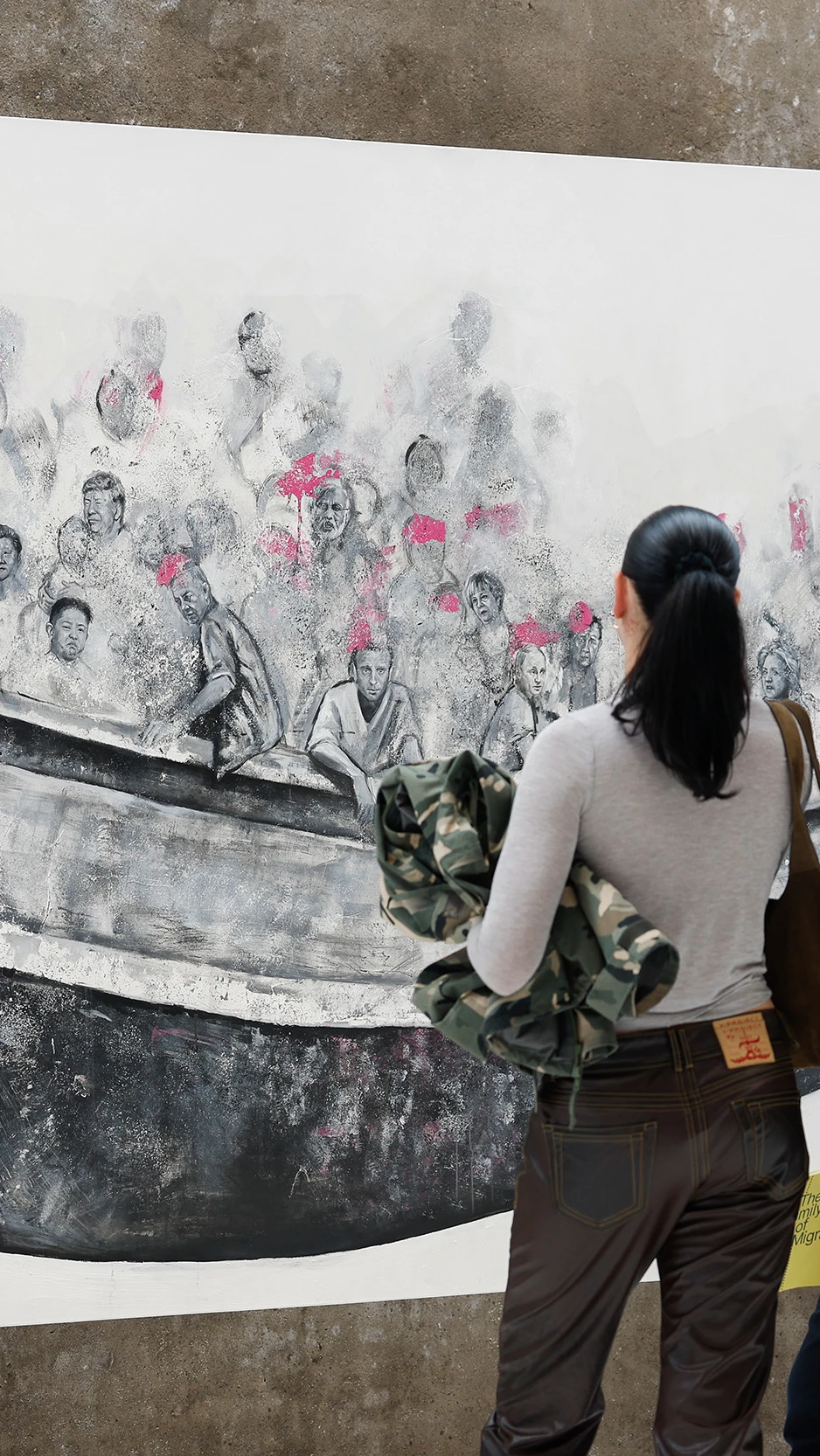 A man and woman observe a large mural of people crowded in a boat, wearing pink life vests. The scen