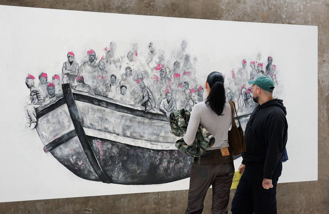 A man and woman observe a large mural of people crowded in a boat, wearing pink life vests. The scen