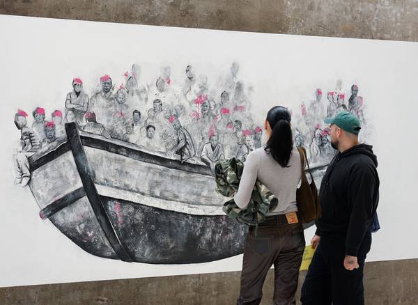 A man and woman observe a large mural of people crowded in a boat, wearing pink life vests. The scen