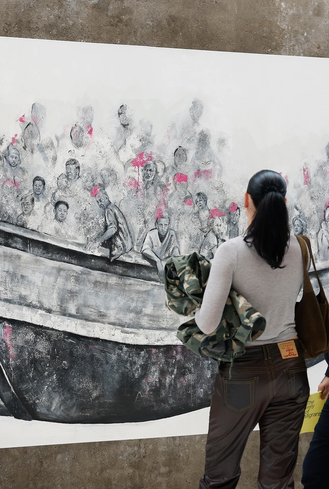 A man and woman observe a large mural of people crowded in a boat, wearing pink life vests. The scen