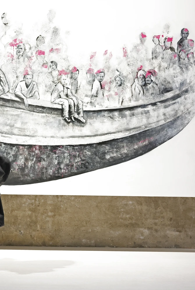 Two people stand in front of a black-and-white mural depicting crowded figures in a boat.