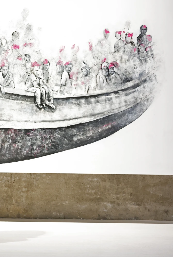 Two people stand in front of a black-and-white mural depicting crowded figures in a boat.