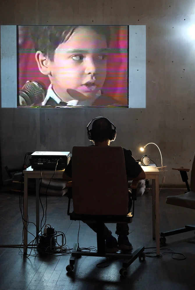 A person wearing headphones sits at a table in a dimly lit room, watching a video projection.