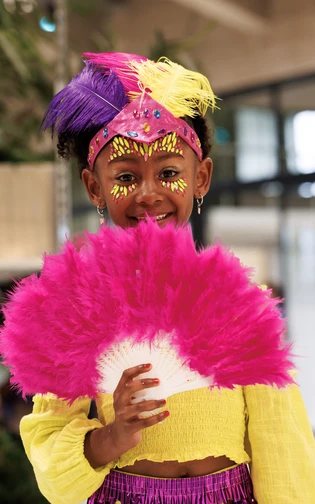 A smiling child in a bright carnival costume holds a large pink feather fan.