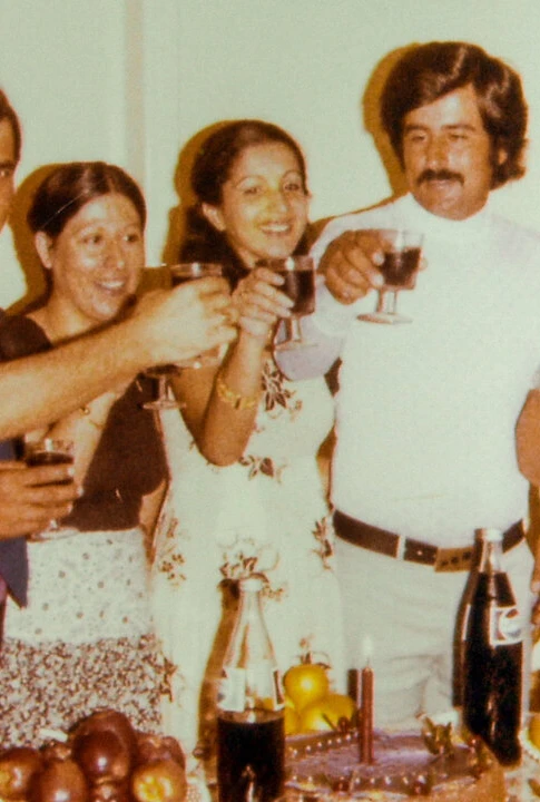 A group of seven people stand closely together indoors, smiling and raising glasses in a toast. A ta