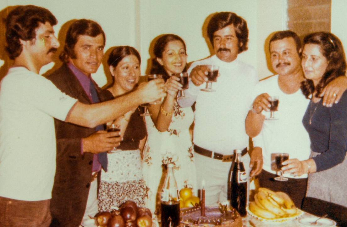 A group of seven people stand closely together indoors, smiling and raising glasses in a toast. A ta