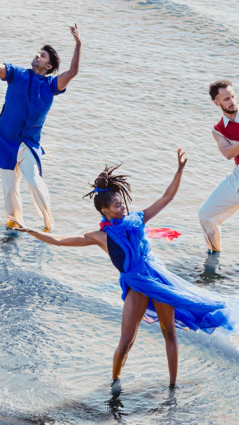 Three dancers perform expressive movements in shallow water, dressed in blue, red and white.