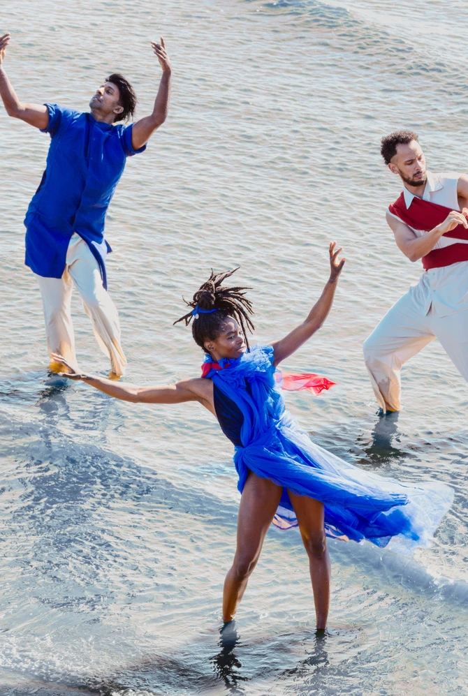 Three dancers perform expressive movements in shallow water, dressed in blue, red and white.
