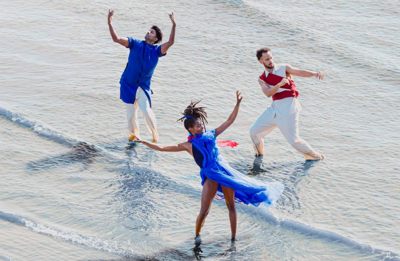 Three dancers perform expressive movements in shallow water, dressed in blue, red and white.