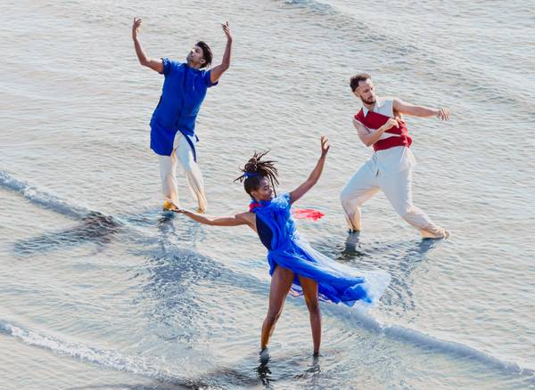 Three dancers perform expressive movements in shallow water, dressed in blue, red and white.