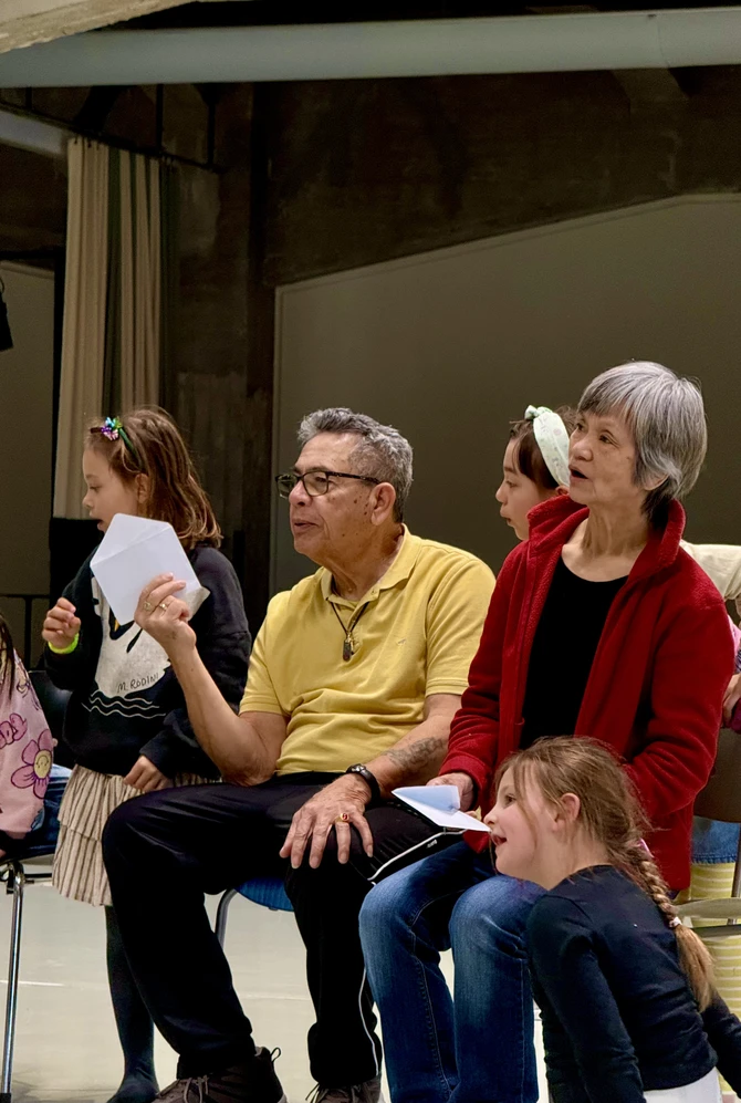 A diverse group of people, including an elderly man and woman, sit attentively with children