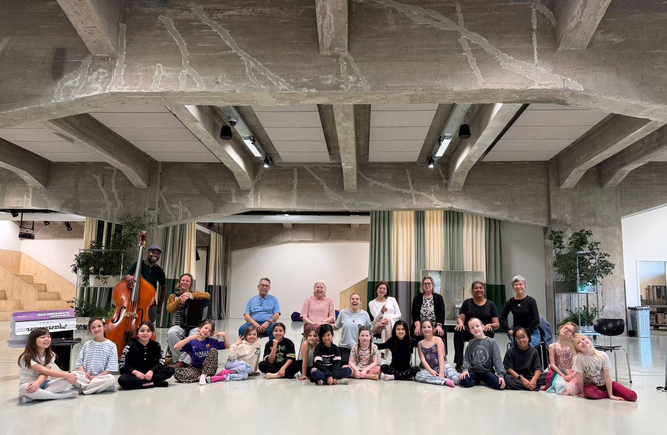 A diverse group of children and adults, some with musical instruments, gather at Plein.