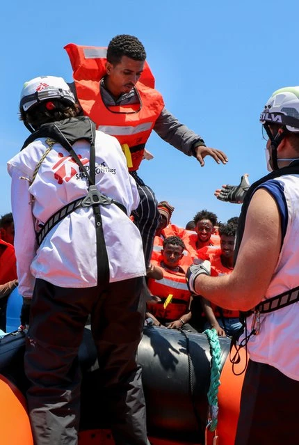 A rescue team assists migrants in orange life jackets on a raft in calm, blue waters.