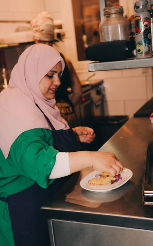 A woman wearing a pink hijab and green top prepares a dish in a kitchen.