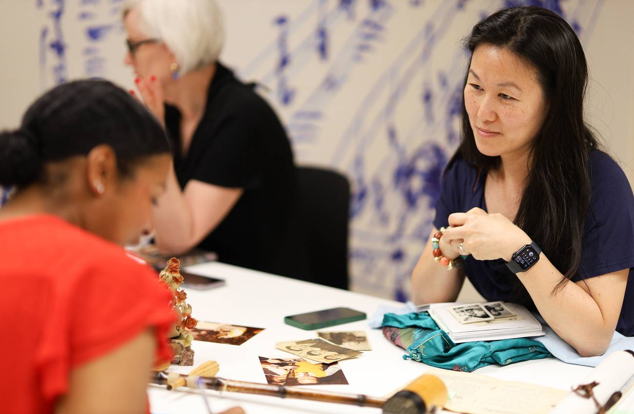 Three women sit at a table engaged in conversation, with photos and items spread out.