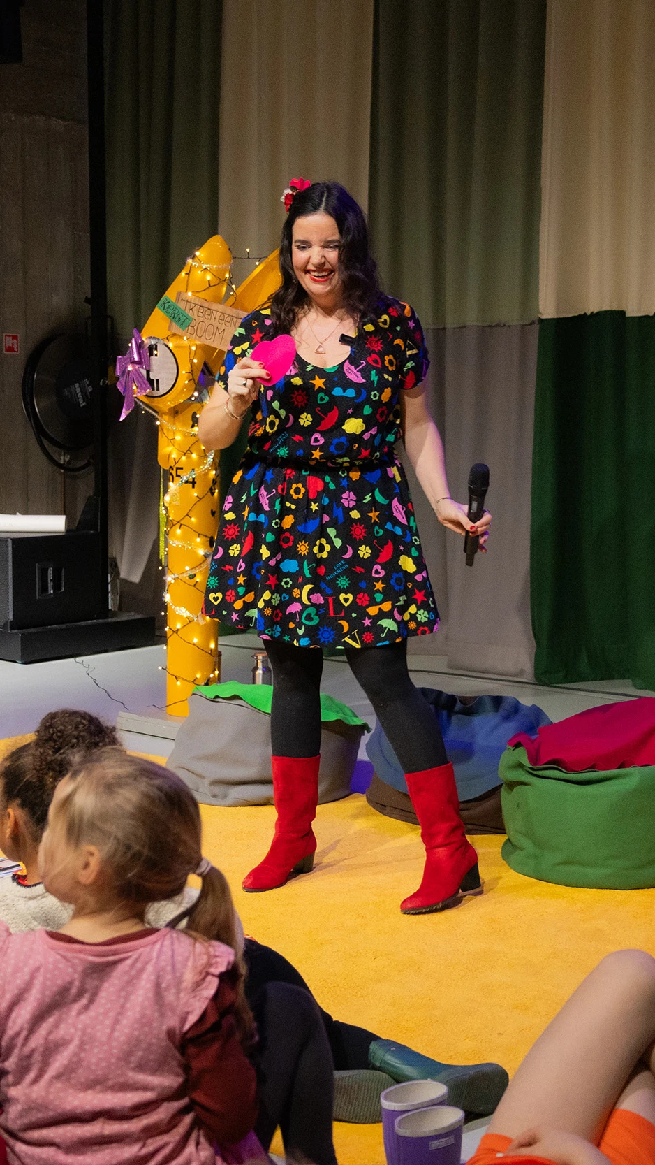 een vrouw in een kleurige jurk staat in een theater voor een groep kinderen op zitzakken