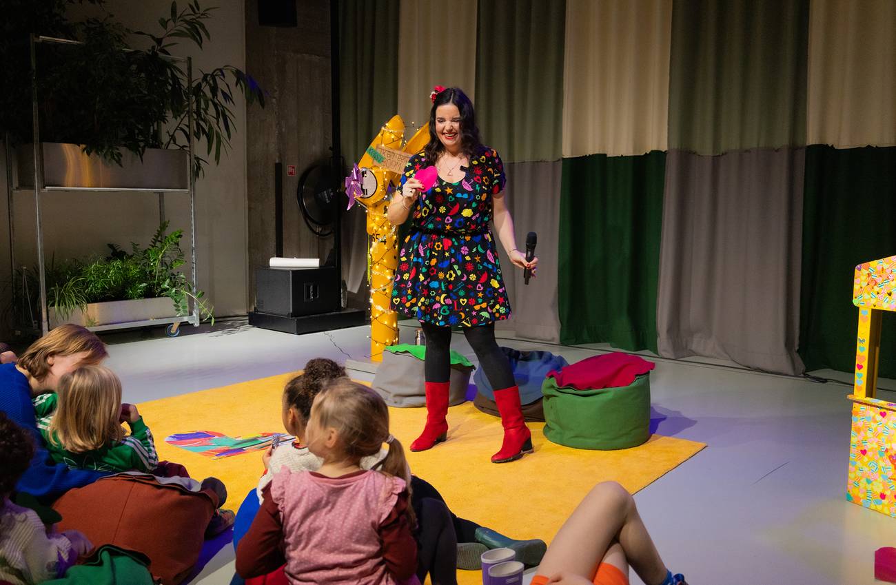 een vrouw in een kleurige jurk staat in een theater voor een groep kinderen op zitzakken