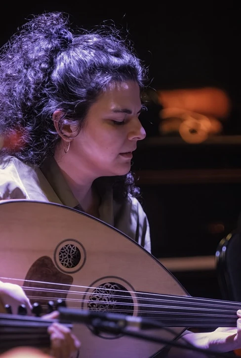 A woman with curly hair plays an oud intently during a music performance. The background is dimly li