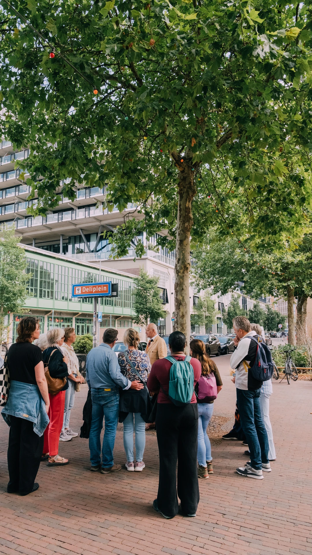 een groepje mensen staat om een verteller heen. op een straatbord staat 'deliplein'