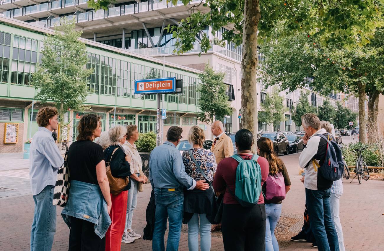 een groepje mensen staat om een verteller heen. op een straatbord staat 'deliplein'