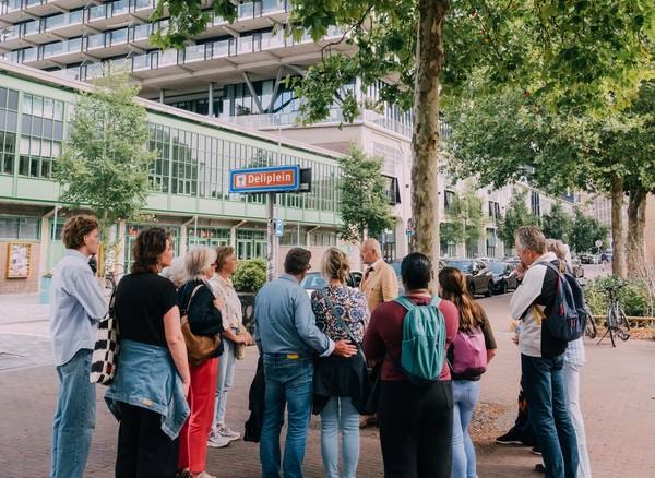 een groepje mensen staat om een verteller heen. op een straatbord staat 'deliplein'