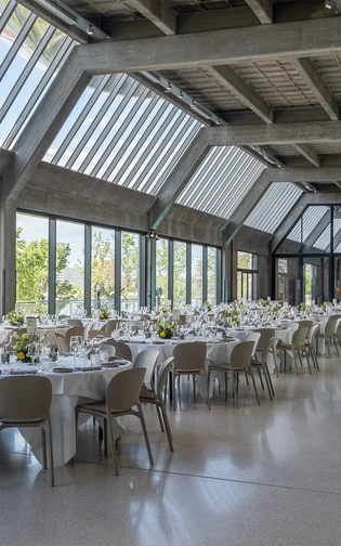 Zaal met ramen van vloer tot plafond met ronde tafels met tafelschikkingen en bloemstukken