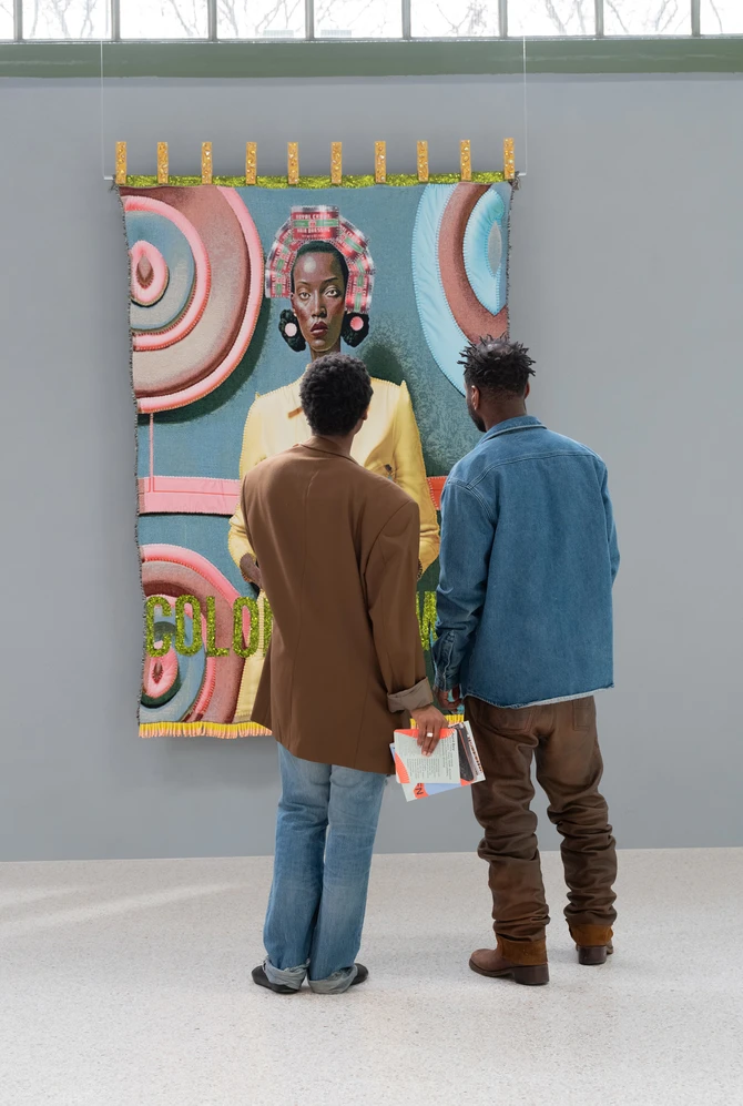Two visitors view a colourful textile portrait displayed in a bright museum gallery.