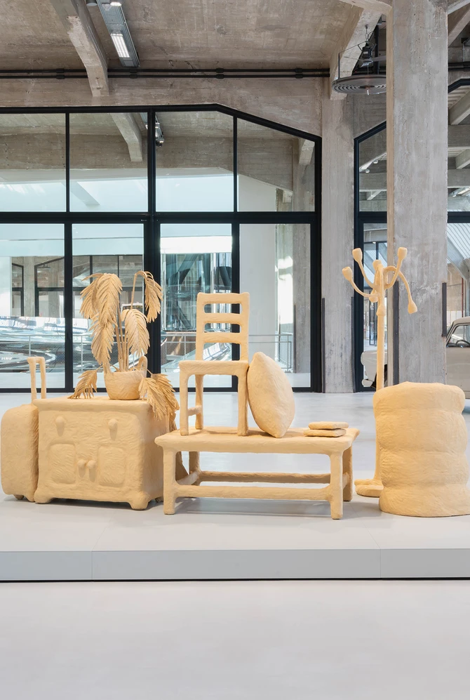 Exhibition space with sculpted furniture on a platform, featuring a chair, table, and coat rack.