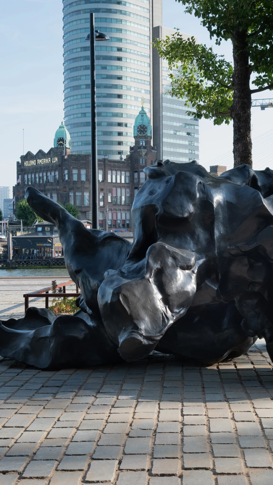 Een abstract bronzen beeld staat op de kade voor Fenix. Achter het beeld is Hotel New York te zien.