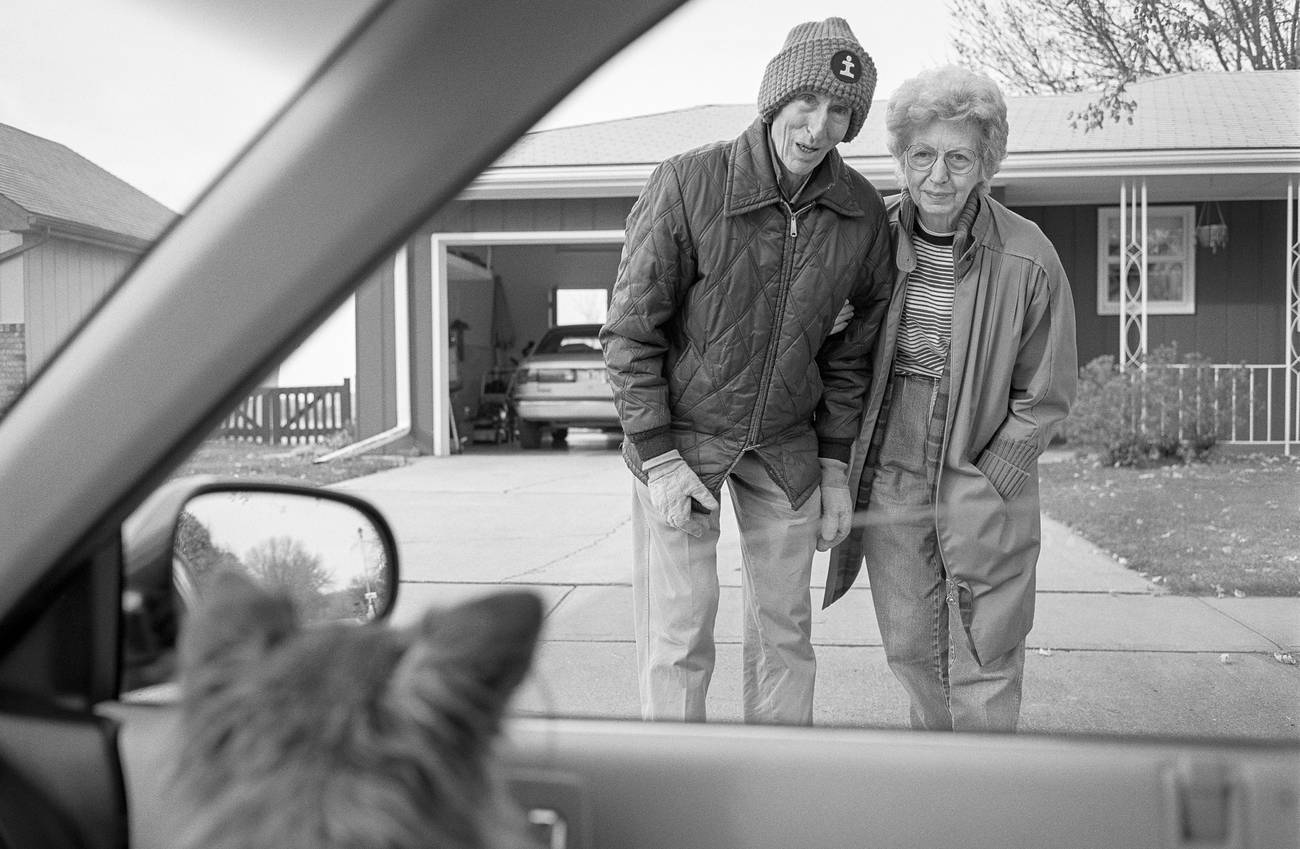 Black-and-white photograph taken from inside a car, showing two people standing in a driveway.