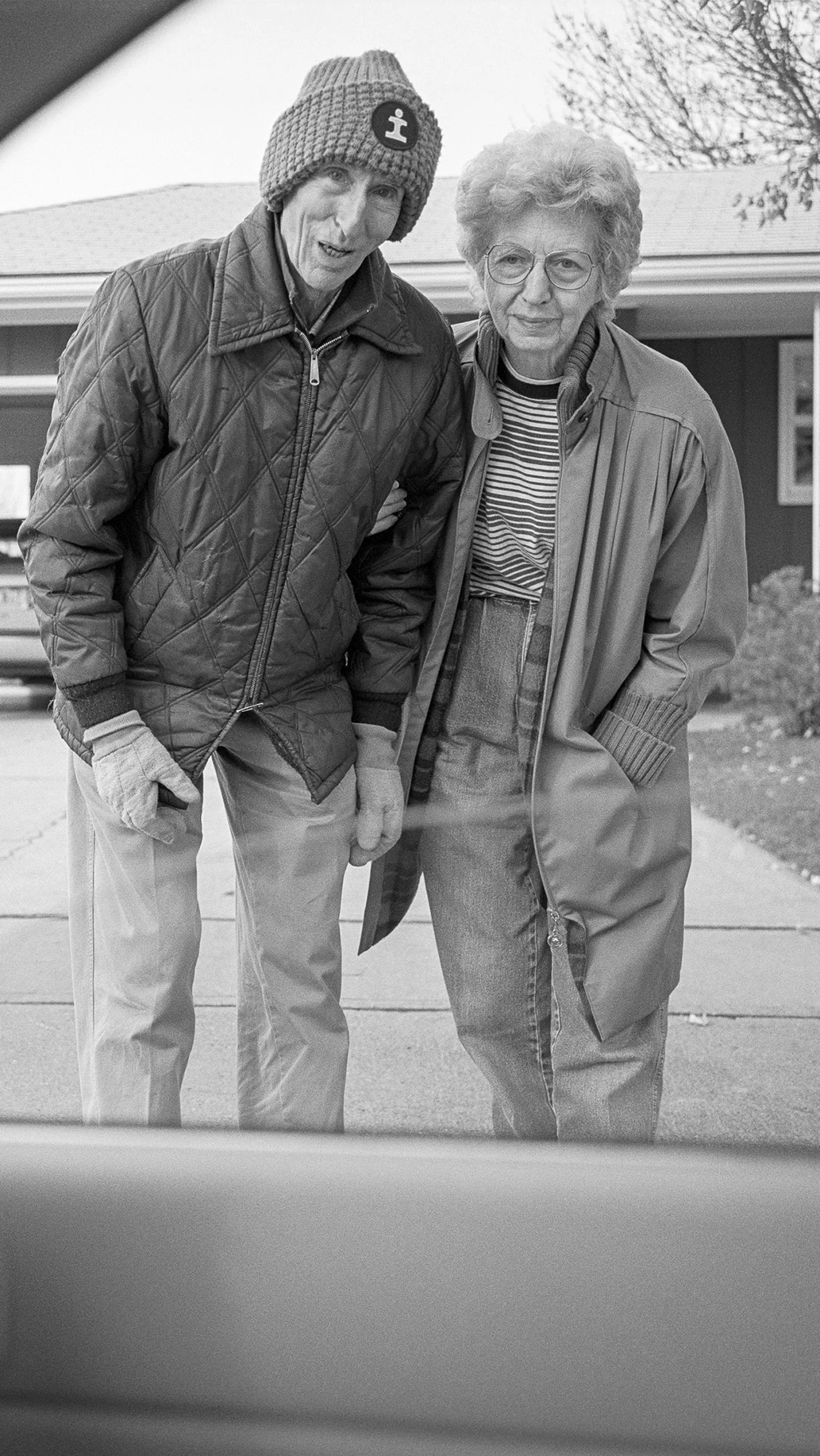 Black-and-white photograph taken from inside a car, showing two people standing in a driveway.