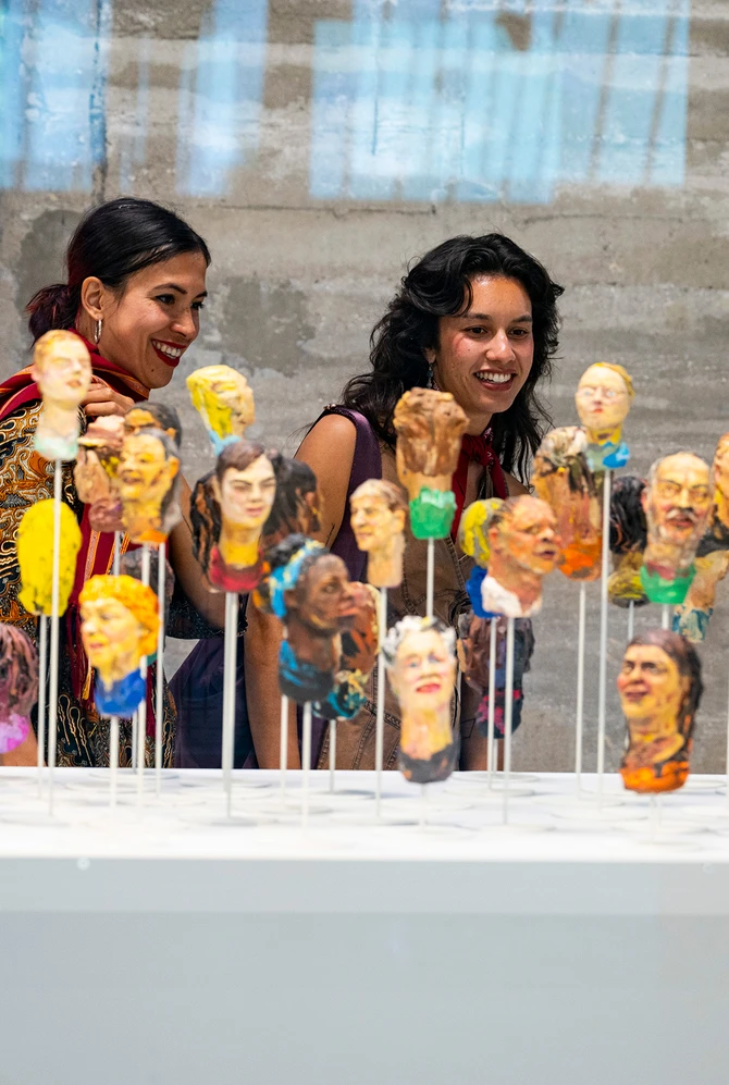 Two women in Fenix admire colorful clay busts on thin sticks. The display is vibrant and diverse