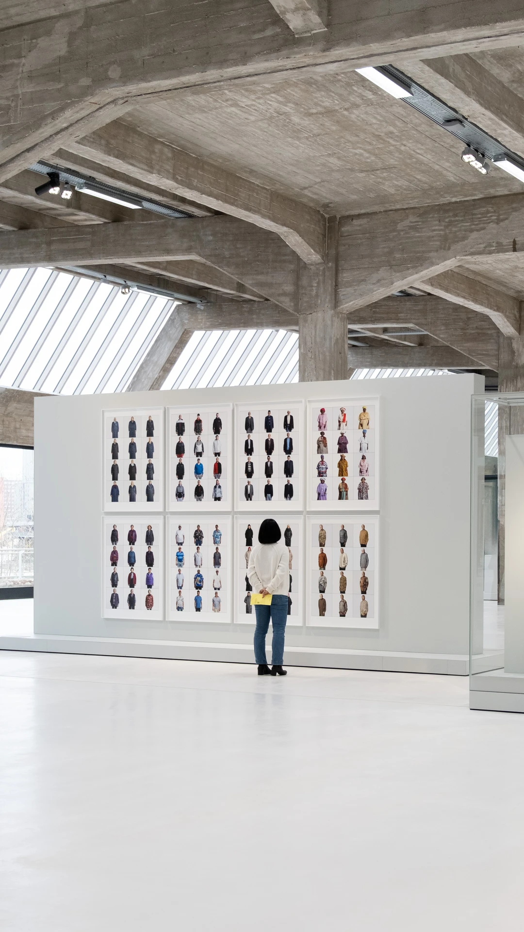 een vrouw staat in een moderne galerie voor grote, witte lijsten met verschillende portretfoto's.