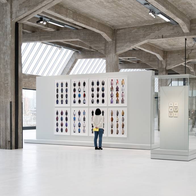 een vrouw staat in een moderne galerie voor grote, witte lijsten met verschillende portretfoto's.