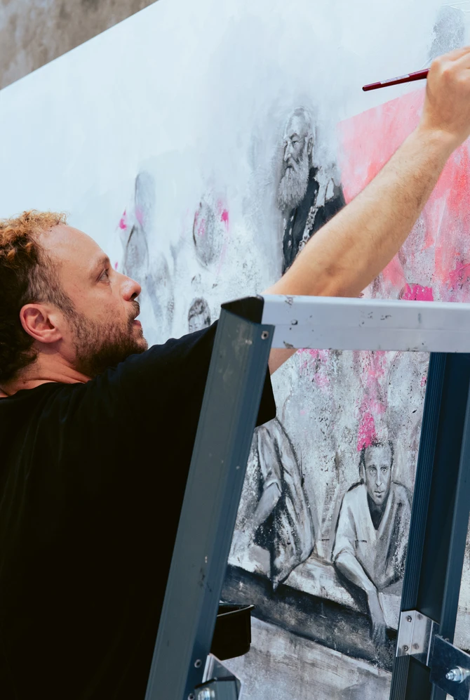 In een kunstgalerie is een man bezig met schilderen op een groot doek, omringd door kunstwerken