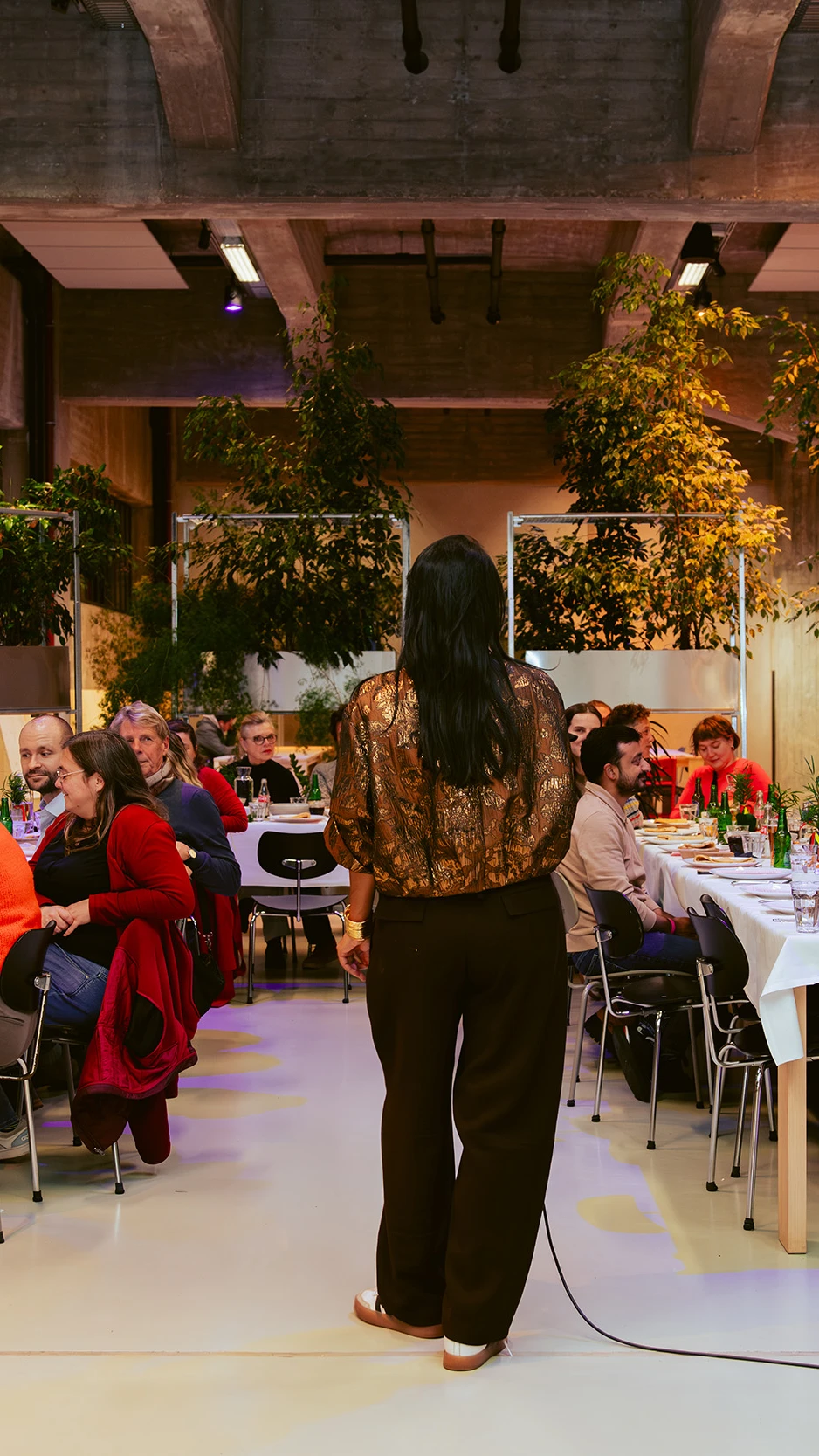 twee lange gedekte tafels met mensen, luisterend naar een vrouw met een microfoon, gezien op de rug