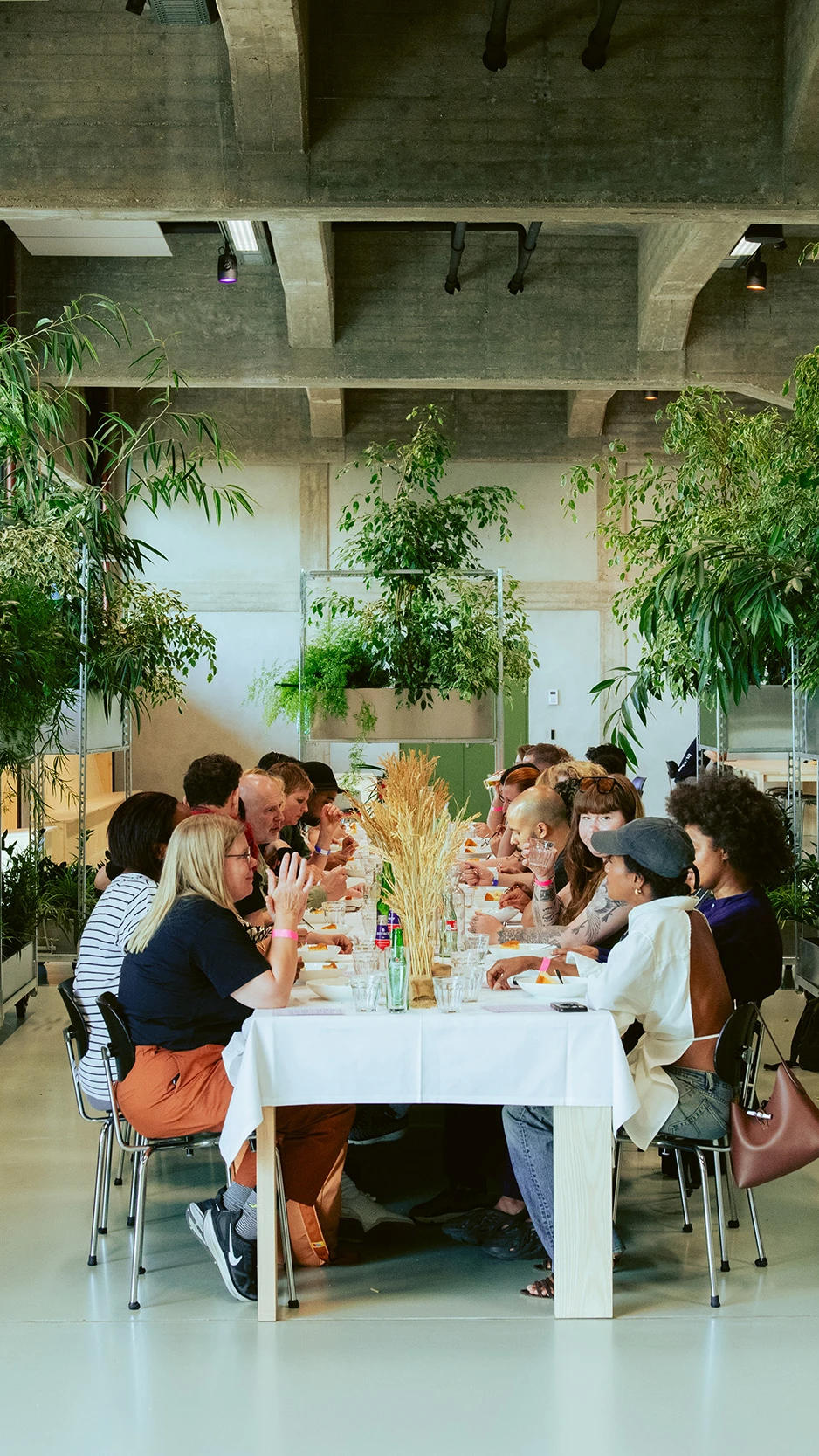 A long dining table with people sharing lunch in a bright space with concrete ceilings and lots of g