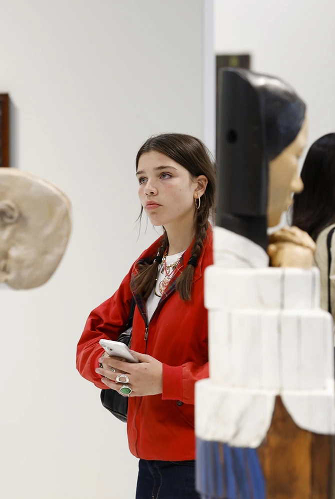 A young woman in a red jacket observes sculptures in an art gallery.