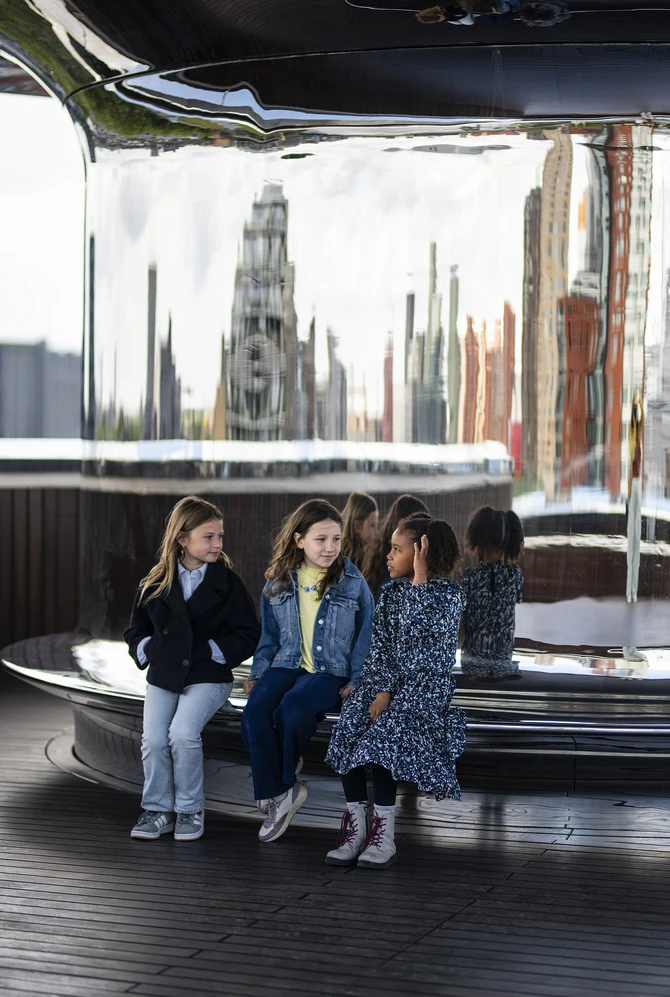 Three children sit together on a shiny, curved bench with reflective surfaces.