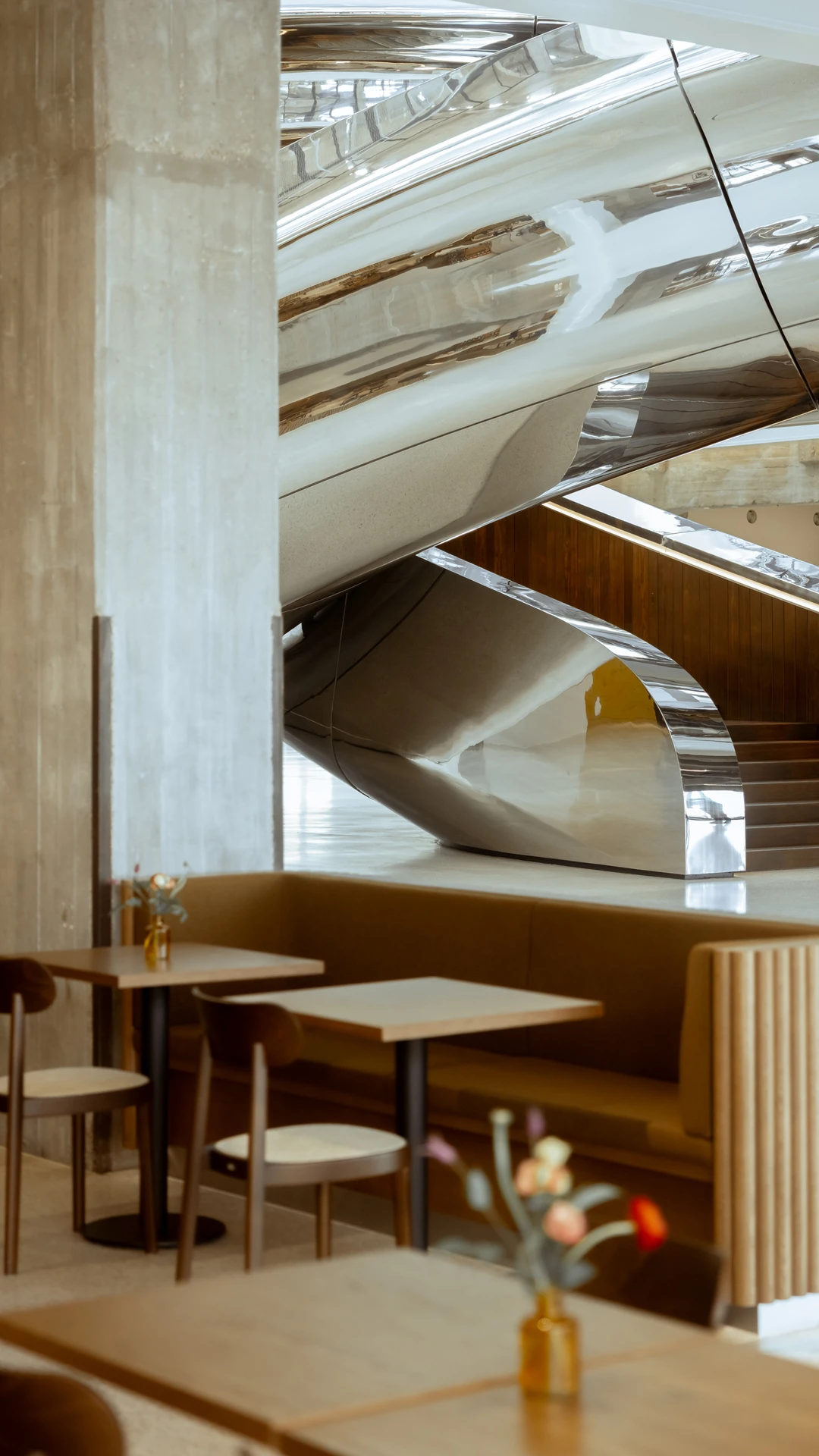 Modern cafe interior with sleek, curved metal stairs, wooden tables, cushioned seating.