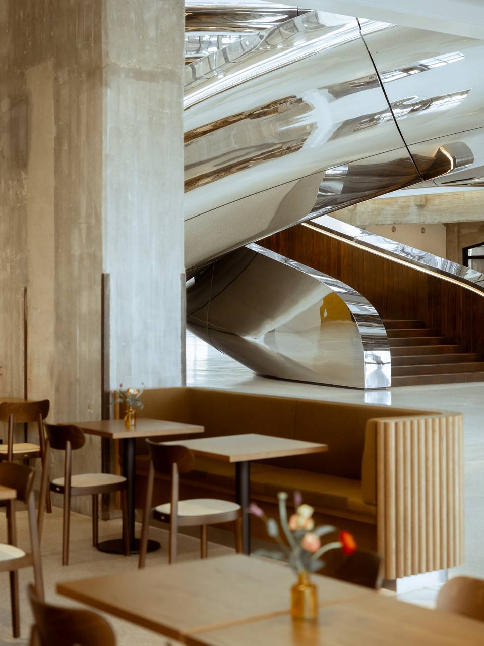 Modern cafe interior with sleek, curved metal stairs, wooden tables, cushioned seating.