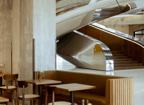 Modern cafe interior with sleek, curved metal stairs, wooden tables, cushioned seating.