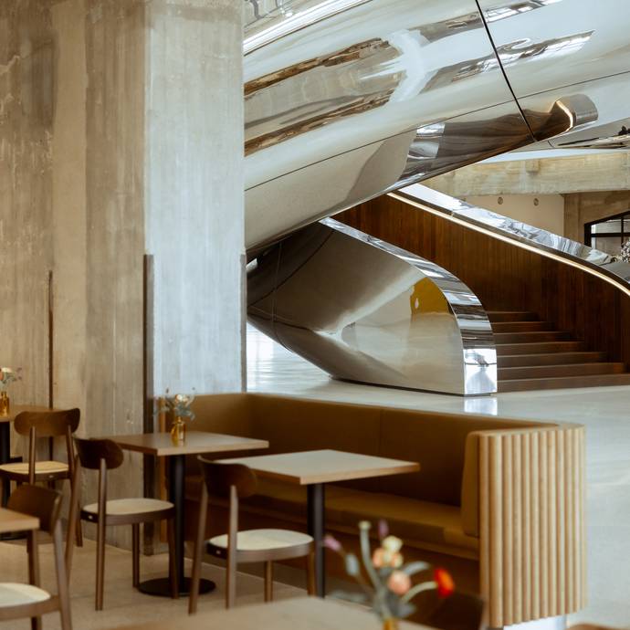 Modern cafe interior with sleek, curved metal stairs, wooden tables, cushioned seating.