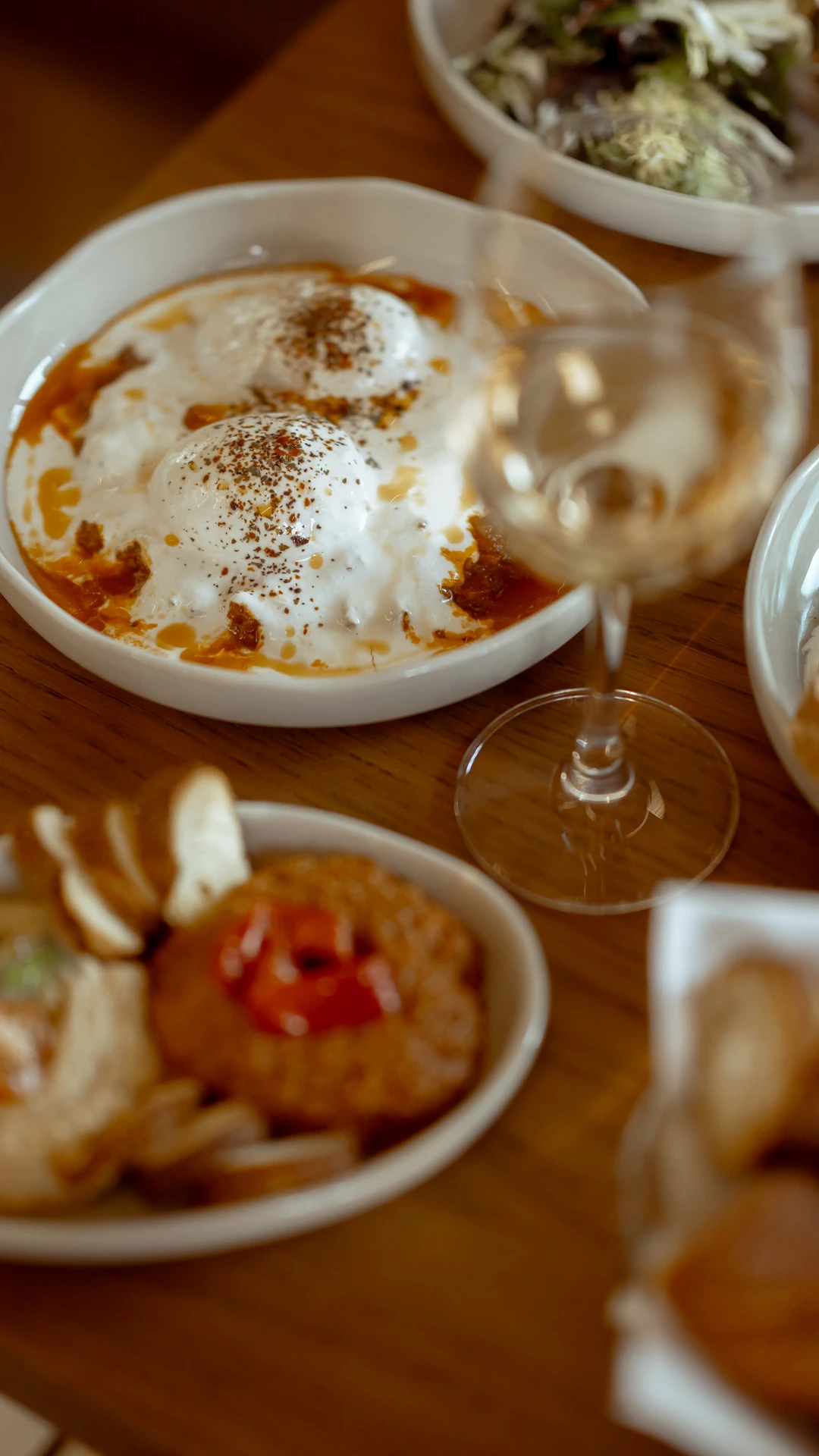 A wooden table with dishes of poached eggs in tomato sauce, salad, bread, dips, and wine.