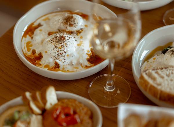 A wooden table with dishes of poached eggs in tomato sauce, salad, bread, dips, and wine.