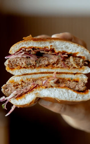 A hand holding a stacked burger cut in half, highlighting layers of juicy beef patty, shredded onion