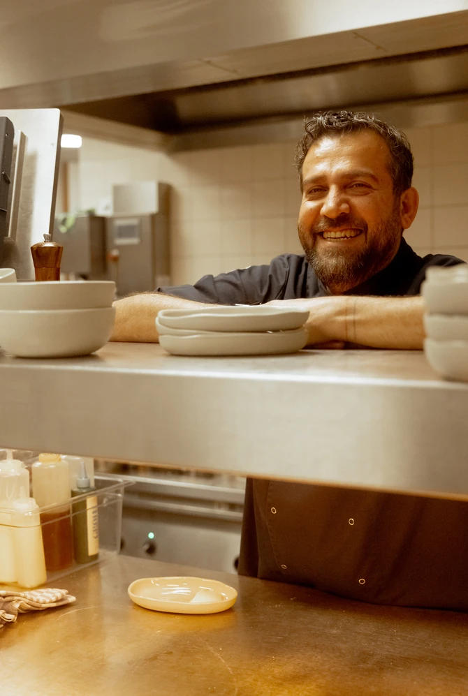 Smiling bearded chef in a kitchen leans on a counter with stacked bowls and plates.